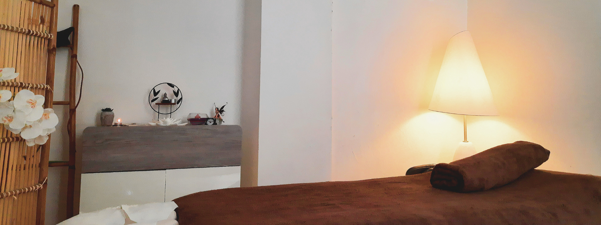 Une jolie lampe éclaire délicatement l'espace bien-être et illumine la table de massages avec douceur en laissant apparaitre une serviette soigneusement roulée et positionnée dessus celle-ci.